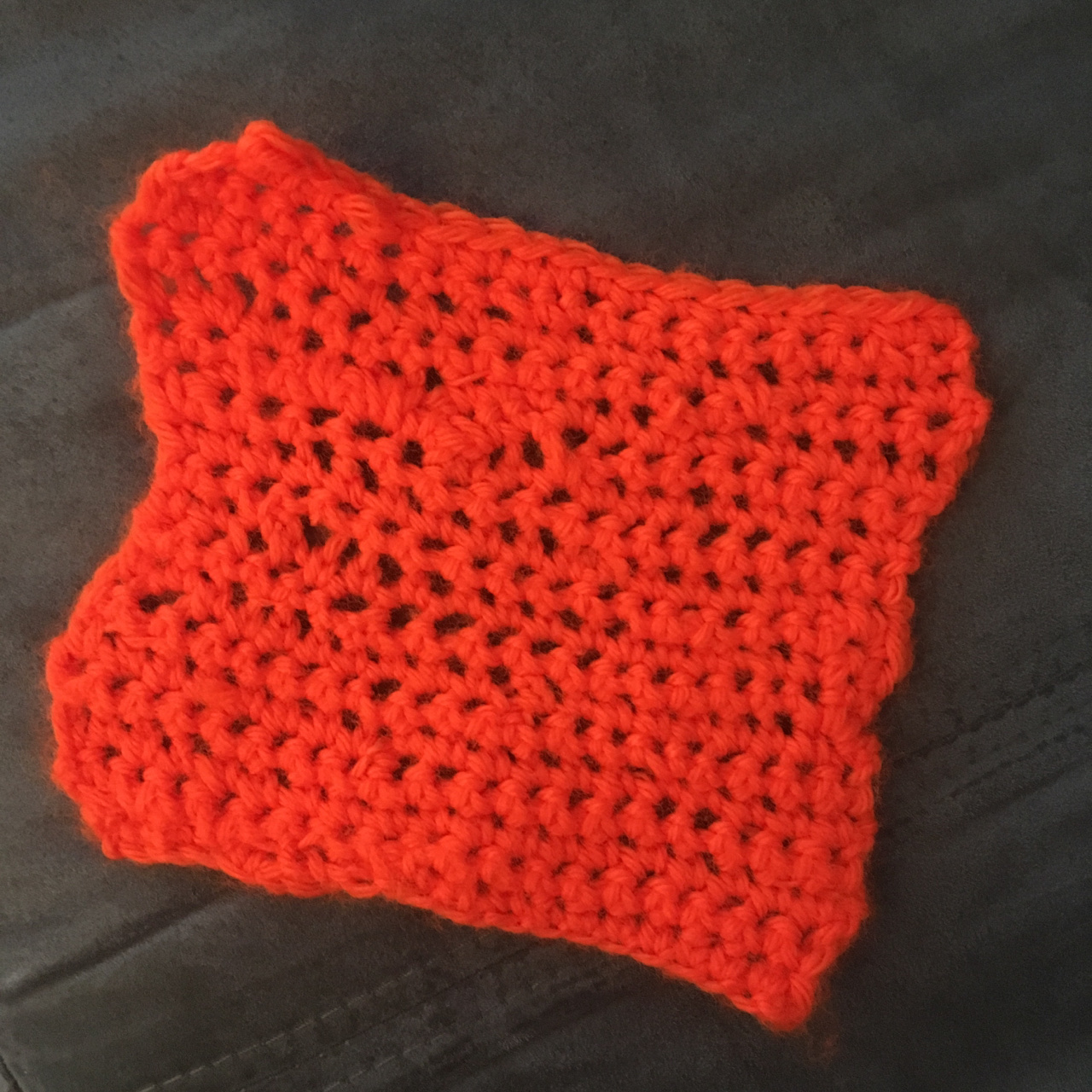 A bright orange dish cloth, wavy around the edges, showing some inconsistency, in stitches, and perfect in its imperfection, lays flat against a grey background.