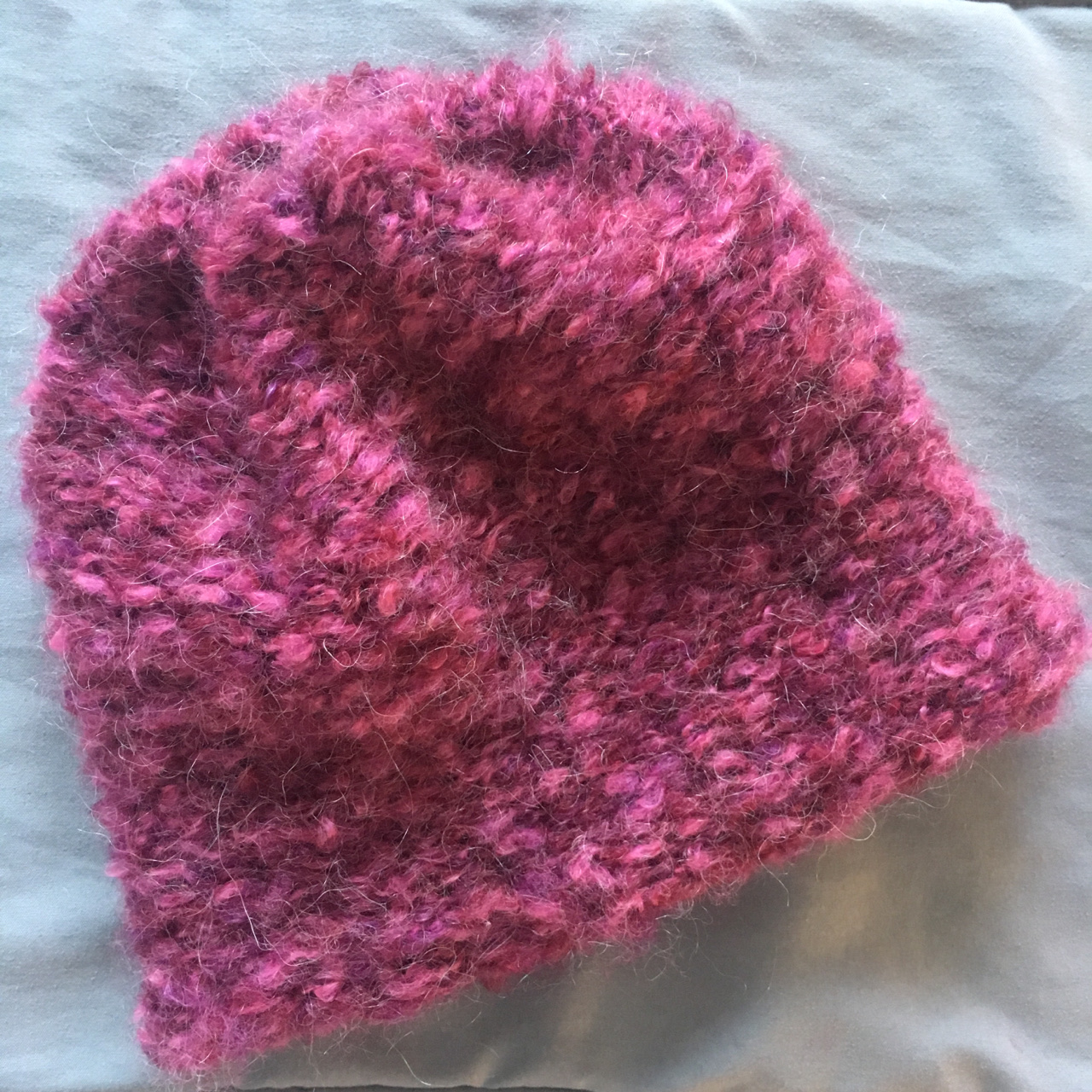 A very simple, fuzzy, brimless beanie in a raspberry purple lays against a grey background.