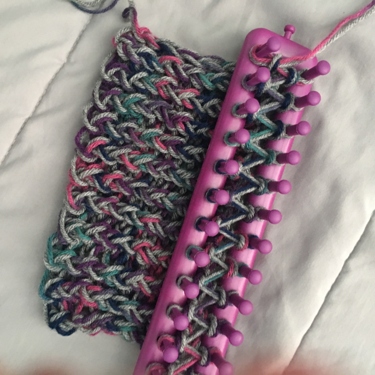 A few inches of thick, squishy scarf double stranded of a varigated green/purple and a smoke grey emerges from a purple 36-peg rake loom against a white background.