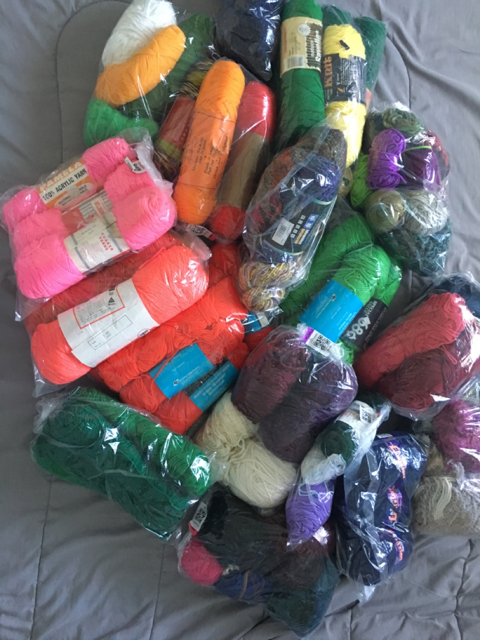 A large and colorful pile of several dozen various skeins and balls of yarn in many colors and textures, atop a grey background.