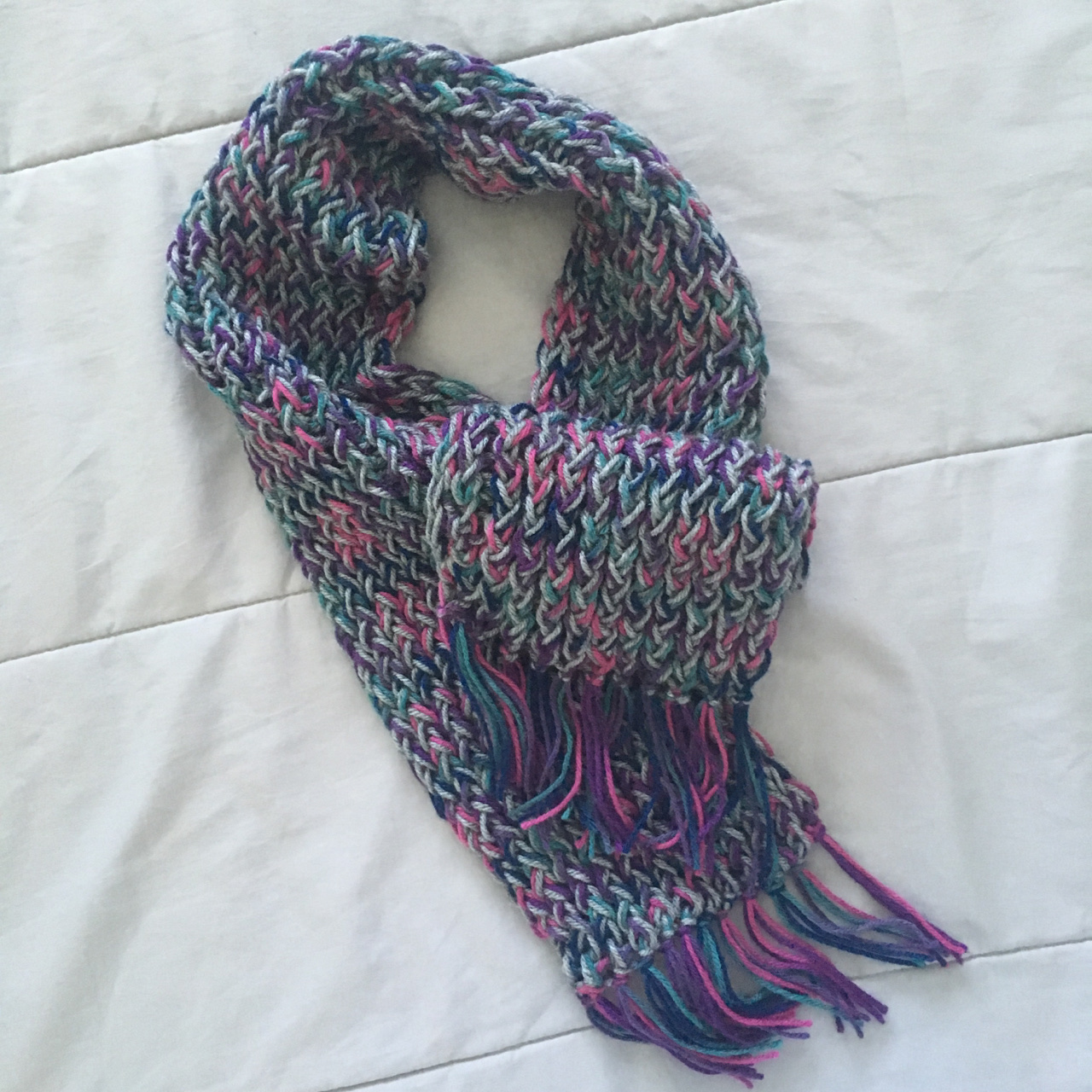 The thick, squishy grey, green, and purple scarf worked up on the rake loom lays decoratively displayed on a white background; fringe has been added to the ends.
