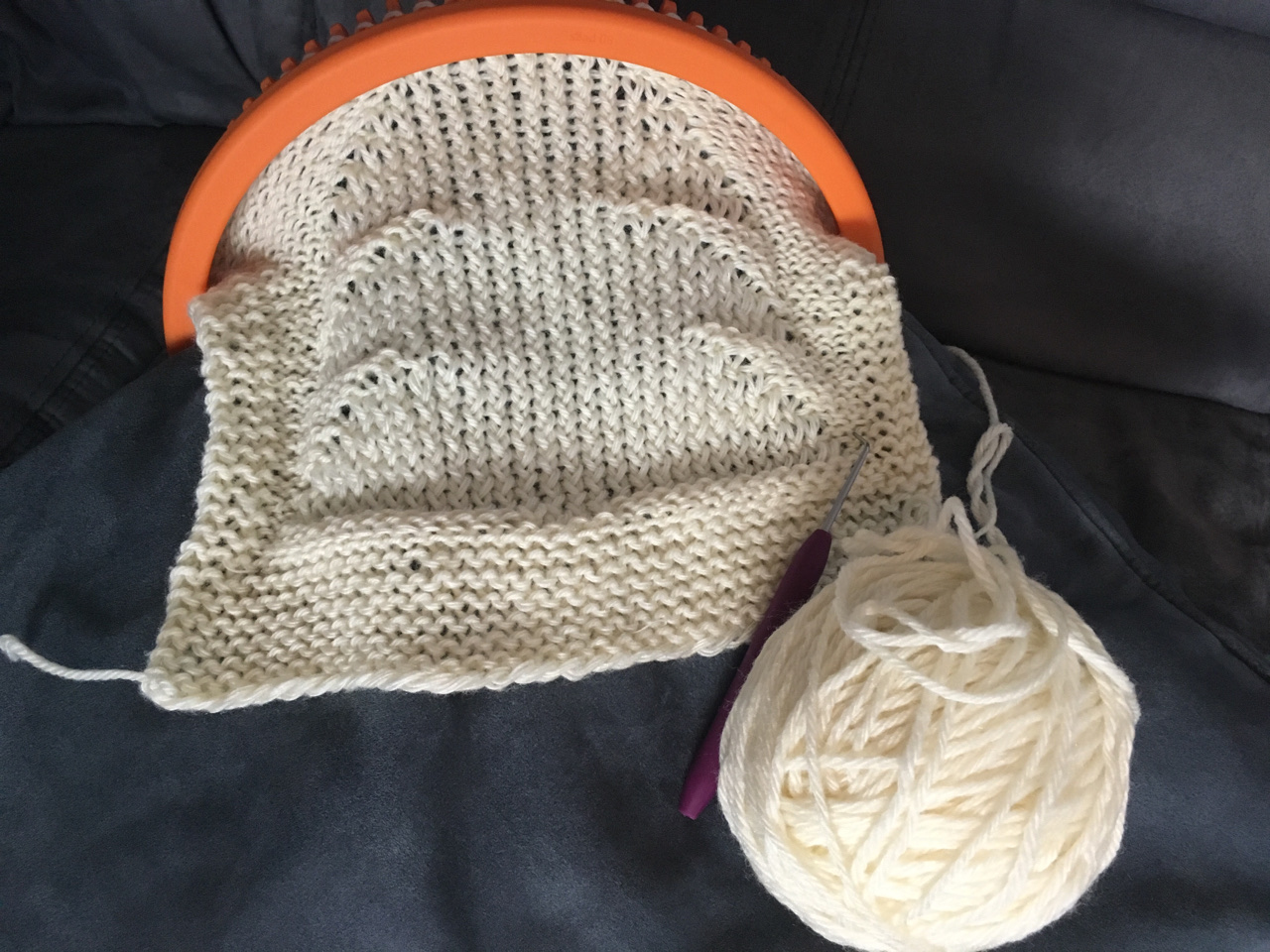 A mostly finished cream colored dish cloth emerges from a bright orange small gauge loo, with an image resembling the shape of a Christmas tree, laid against a dark eggplant colored background.