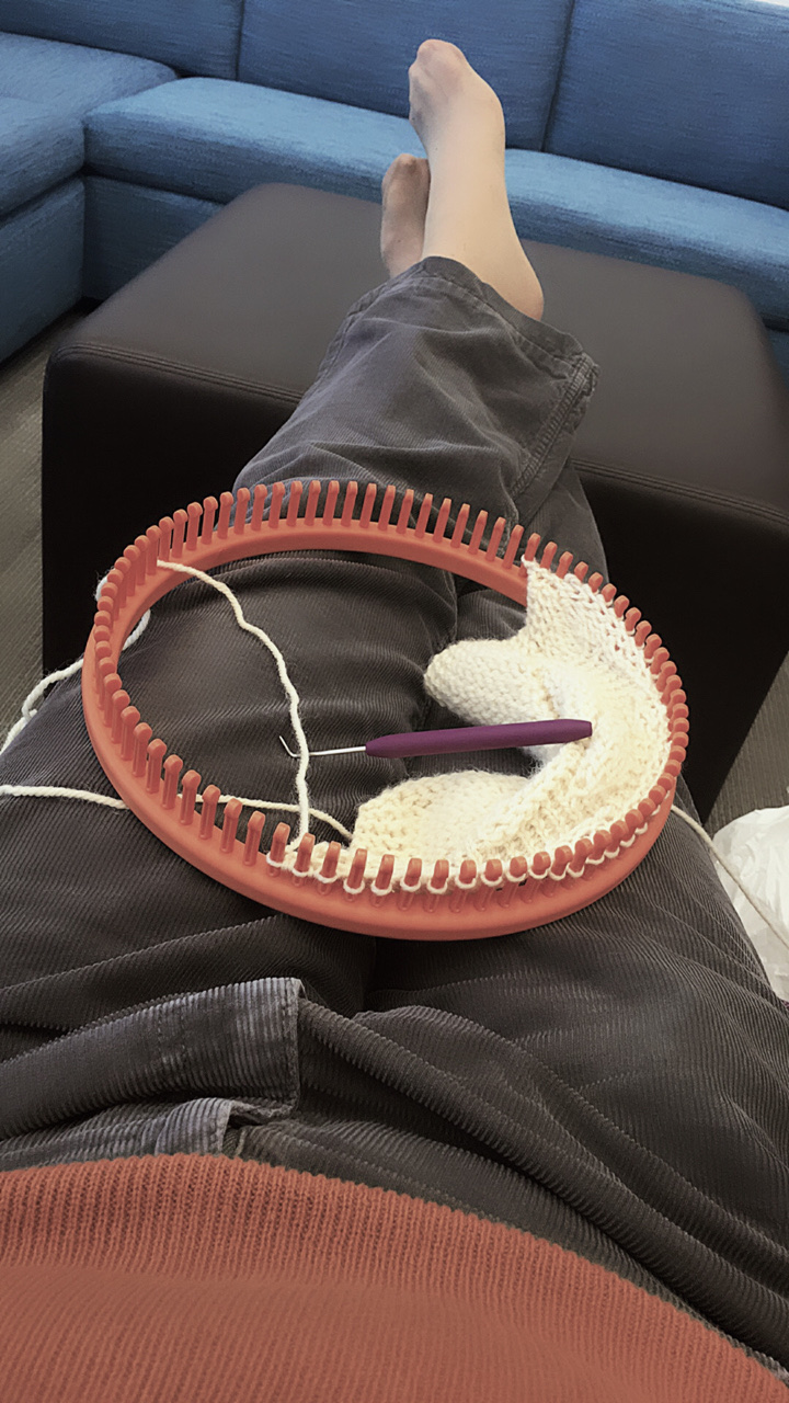 POV my vantage point as reclined in a chair. My feet outstretched and up on a table/footrest. My orange shirt is visible at the bottom edge of the image and I’m wearing grey courderoys and white socks. A bright orange 80-peg small gauge loom sits in my lap, with the Halloween dish cloth emerging from it.