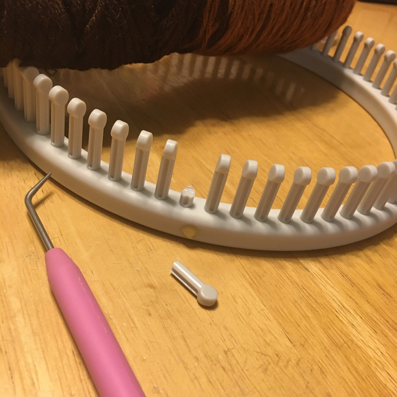 A greyish white 72-peg small gauge loom sits on a light woodgrain tabletop, a skein of dark brown yarn sits atop it in the background. At the front of the loom in the foreground, a broken peg lays sadly on its side in front of the gap-toothed place on the loom from which it came. A loom tool lays in the foreground to the left.