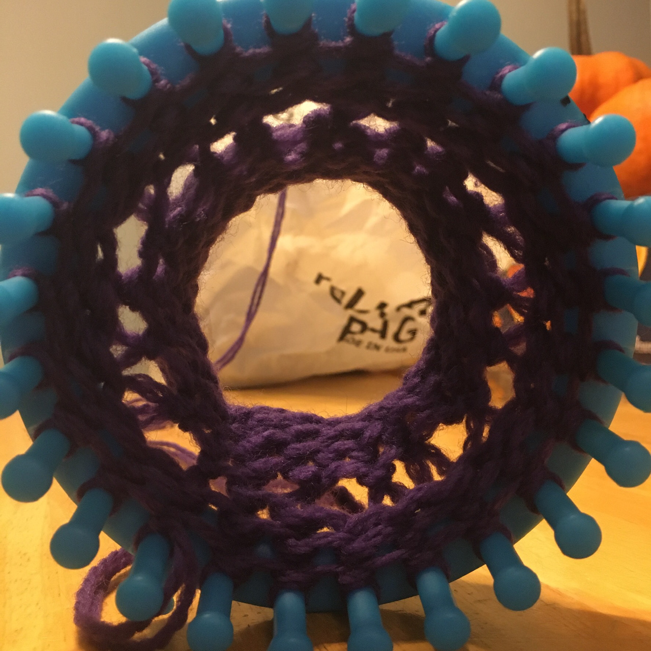 POV looking through the center of a bright blue 24-peg loom standing upright on its side, royal purple yarn growing out the back with a series of intentional holes ringing the work.