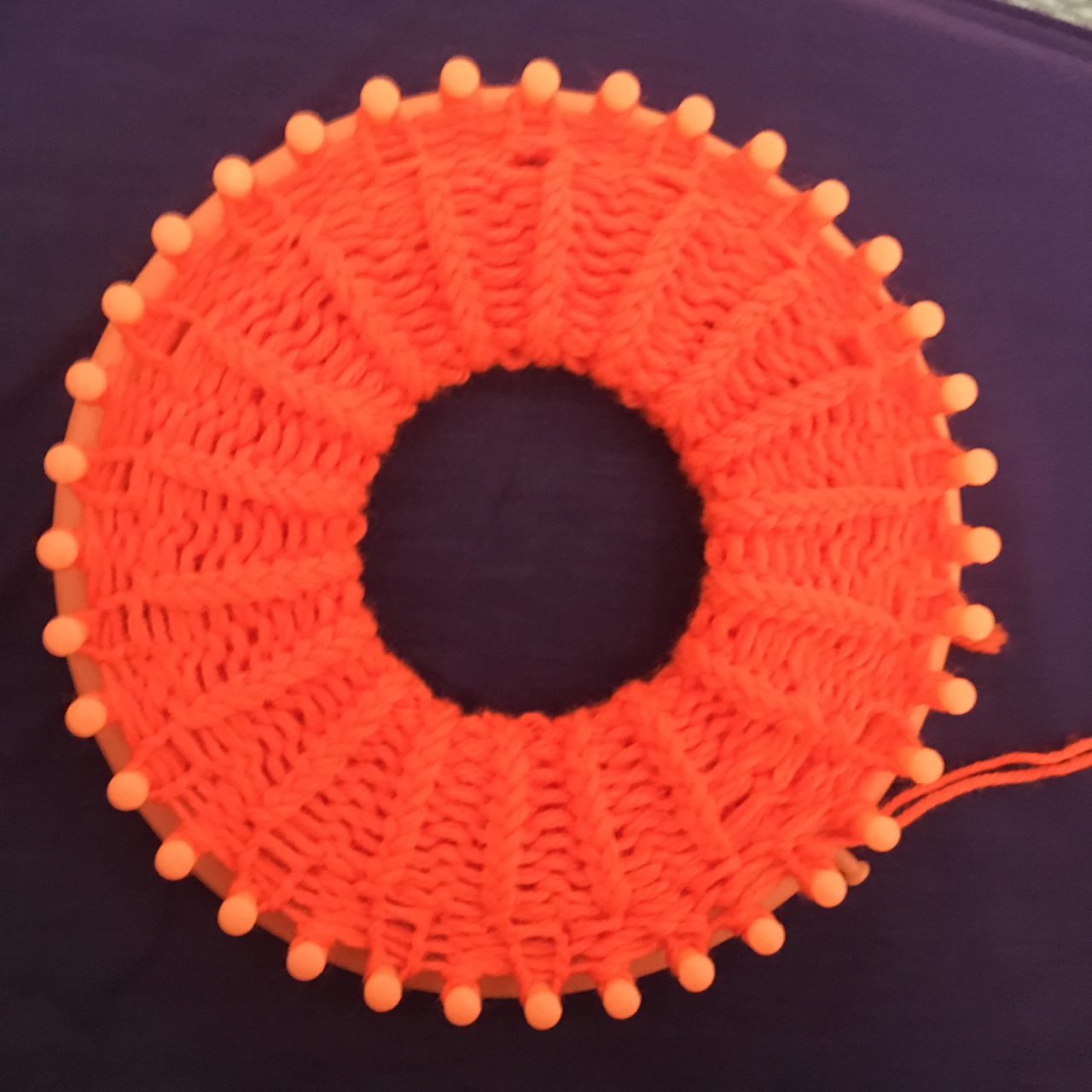 Same bright orange 36-peg loom holding several rows of stitches of the bright orange yarn, the makings of most of a brim, that are stretched tight reaching toward the center of the loom, resembling a spider web, set against a dark eggplant colored background.