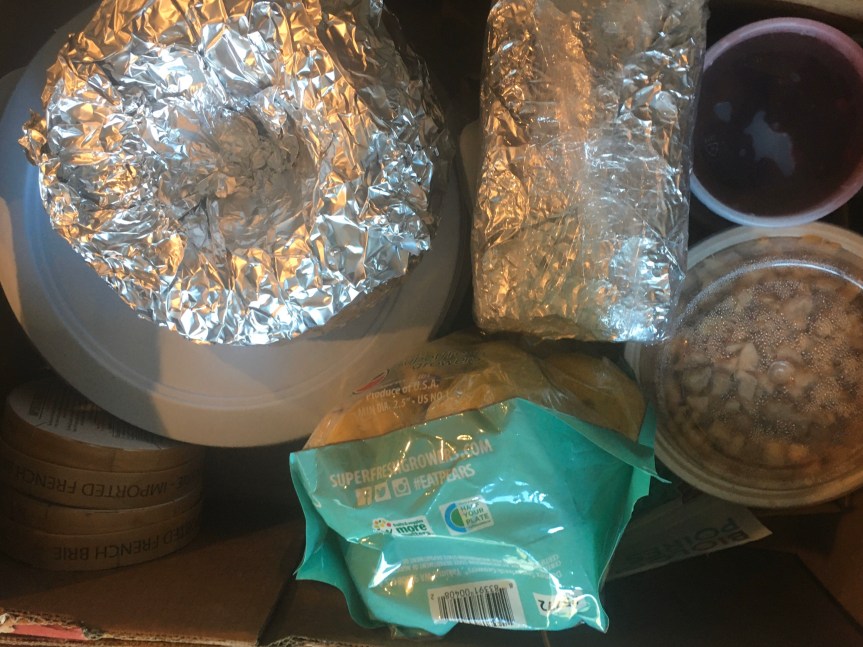 Aerial view of a sturdy cardboard box filled with ingredients and finished products to contribute to a Thanksgiving feast—a bundt cake and loaf, wrapped in foil, wheels of brie, a bag of pears, a tub of cranberry sauce, a pound of bacon, and other food items.