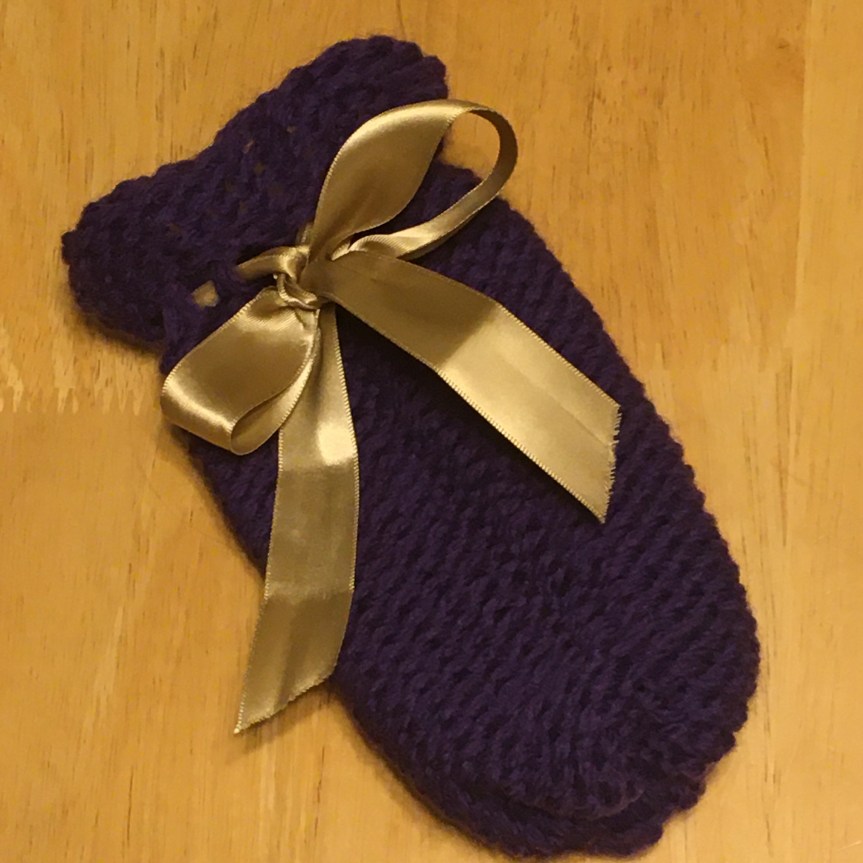 A small, royal purple gift bag lays flat on a light woodgrain background, with a shiny gold ribbon woven through it top as a drawstring and tied in a bow.