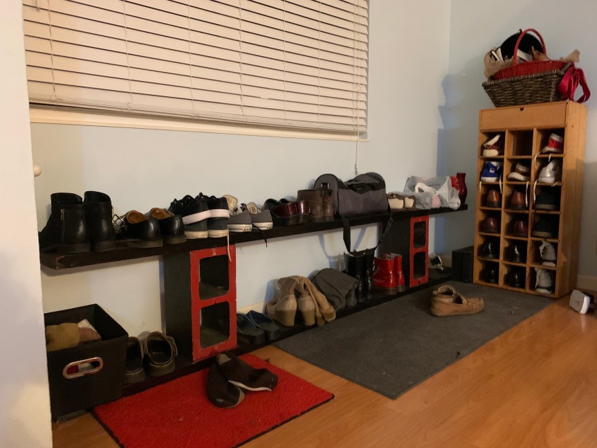 View, from a slight angle, of low brick and board shelves painted black and red, two-tiered and about six feet long, lined with a variety of shoes. Next to it stands a blonde wood shoe cubby, about four feet high and two feet wide, with 18 cubby holes filled with shoes.