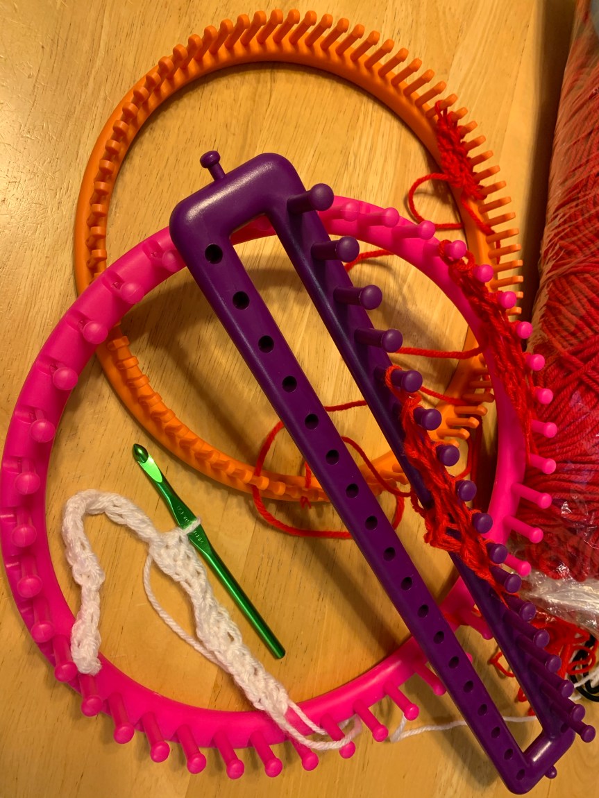 Several different looms piled atop one another, each with the same corner to corner project started in red yarn. Inside one loom sits a small sample swatch of white yarn with a crochet hook holding a loop mid-row working double crochets.