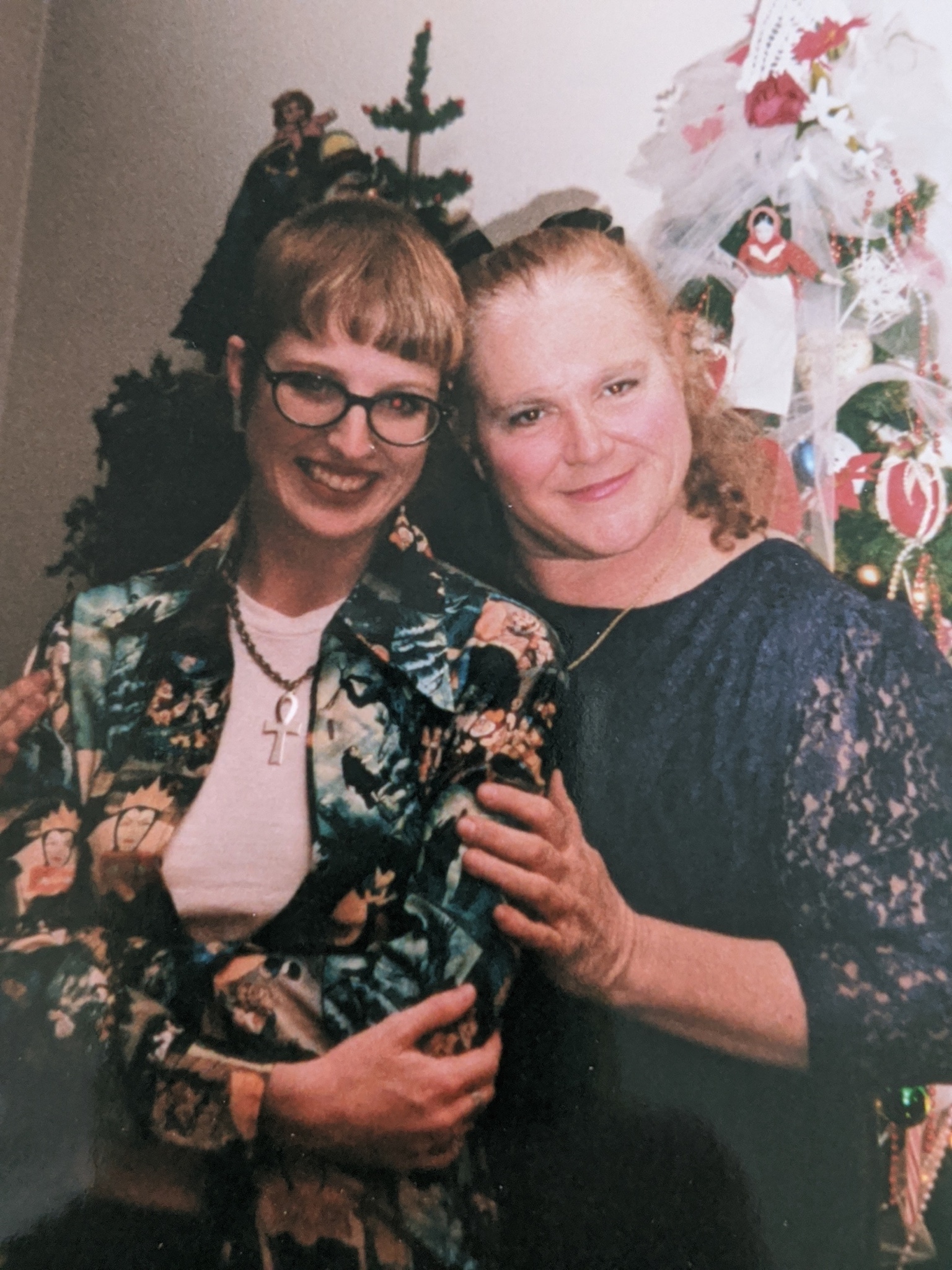 A young Serena, left, with pixie-short red hair, thick black framed glasses, and a casual, funky style of dress, poses for a holiday picture in front of a Christmas tree with her mother, right, smiling serenely and dressed in holiday casual. They lean in gently toward one another.