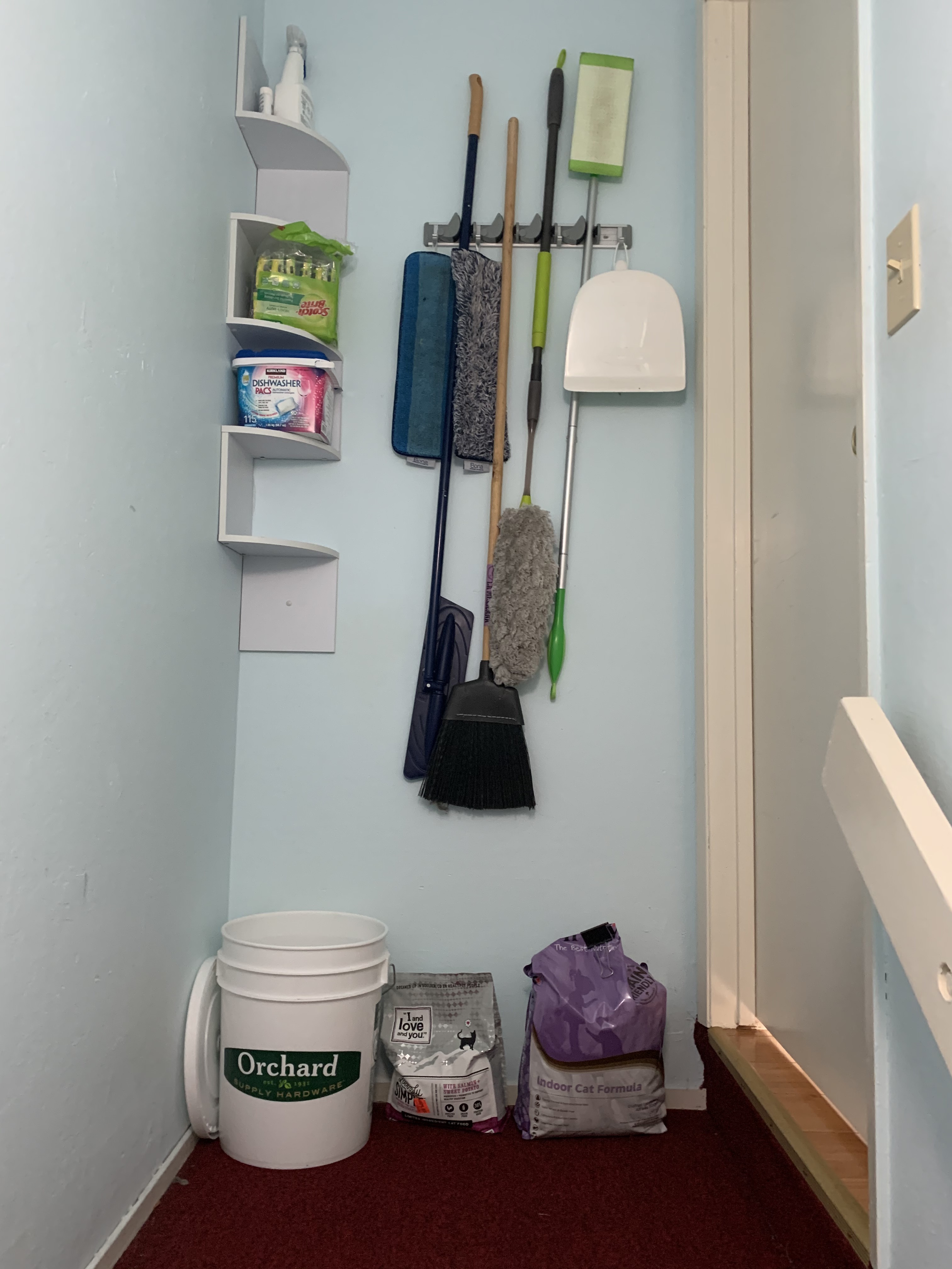 View of our stairwell landing from a few stairs down, looking up. A small set of corner shelves is mounted in the left corner holding a few small cleaning supplies, and a rack to hold brooms and mops is mounted on the wall to the right of it, holding brooms, mops, Swiffer, and a nesting dustpan/brush combo. On the floor in the left corner stands a large white bucket for rags and kitchen towels, and two small/medium sized bags of cat food lean against the wall to its right.