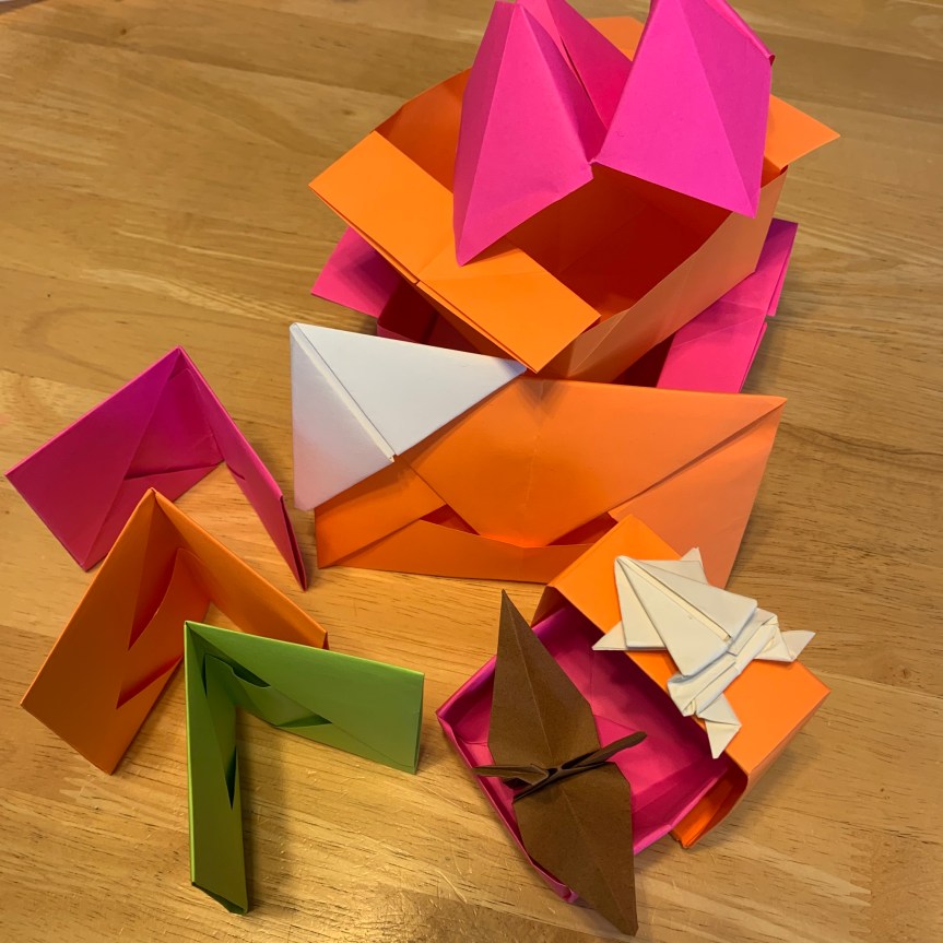 A collection of various origami models arranged together on a tabletop in bright orange, pink, and green. There are wallets standing in a row, boxes and envelopes, topped with a crane and a frog, a bookmark rests at the corner of an envelope, and a fortune teller props up from behind.