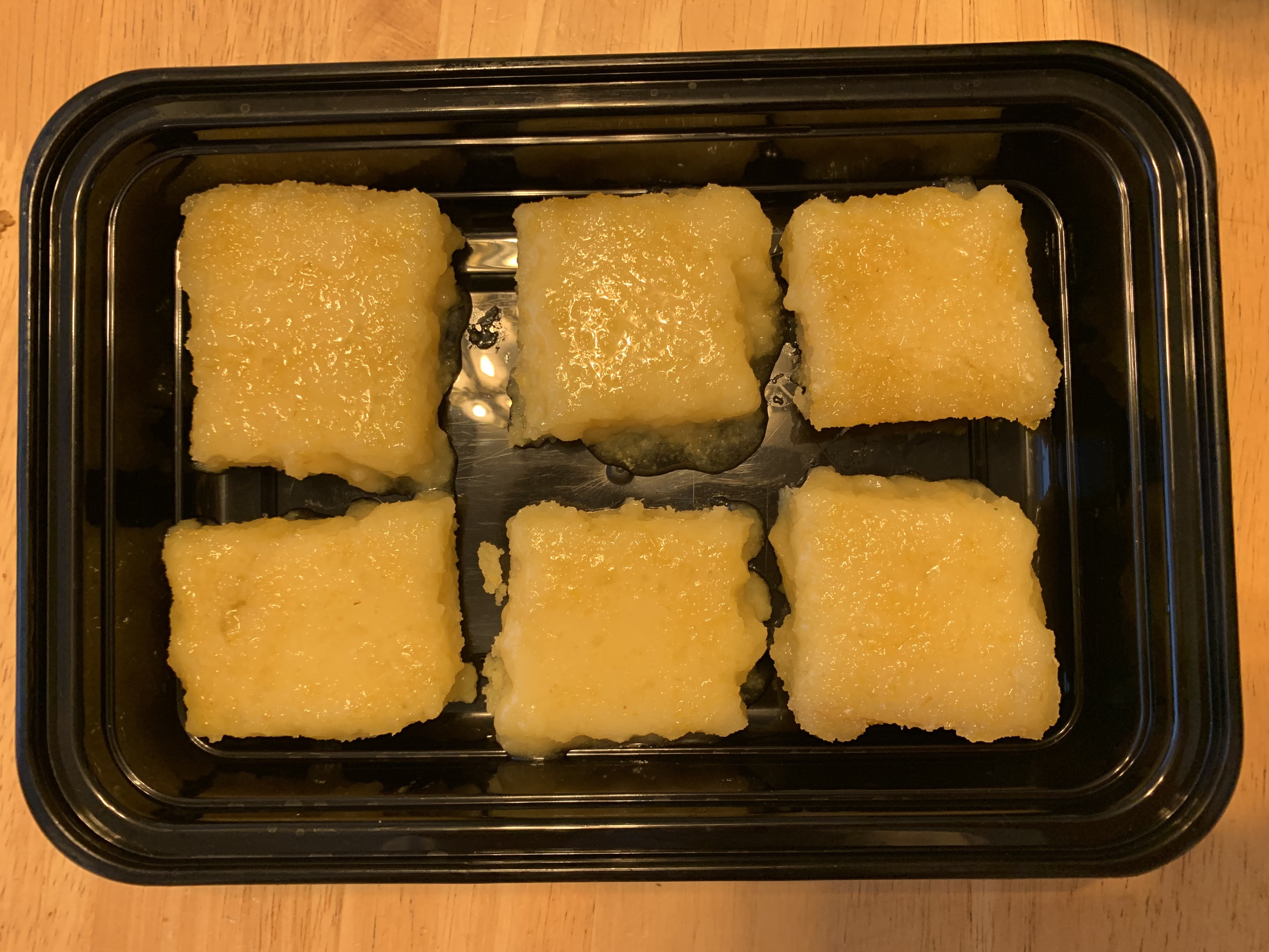 Landscape orientation of a black, rectangular food container holding six bright yellow dessert squares.