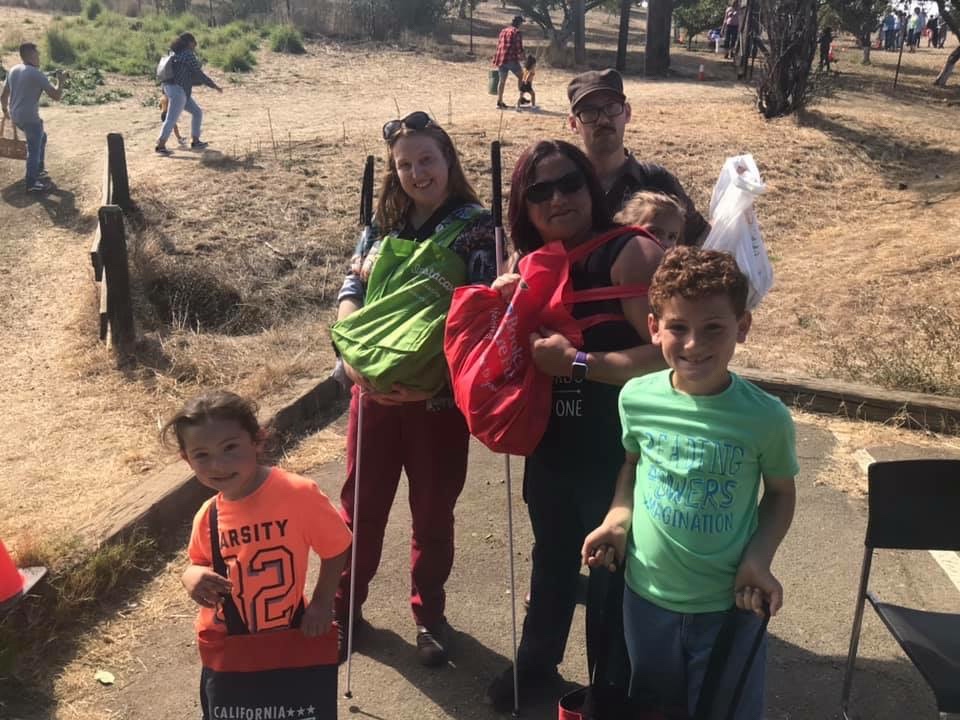 Serena, Sterling, Bay Area Blind Mom, and her three kiddos, the smallest in a carrier on her back, stand together on a dry, grassy hillside near the entrance to the orchard, holding large bags full of apples.
