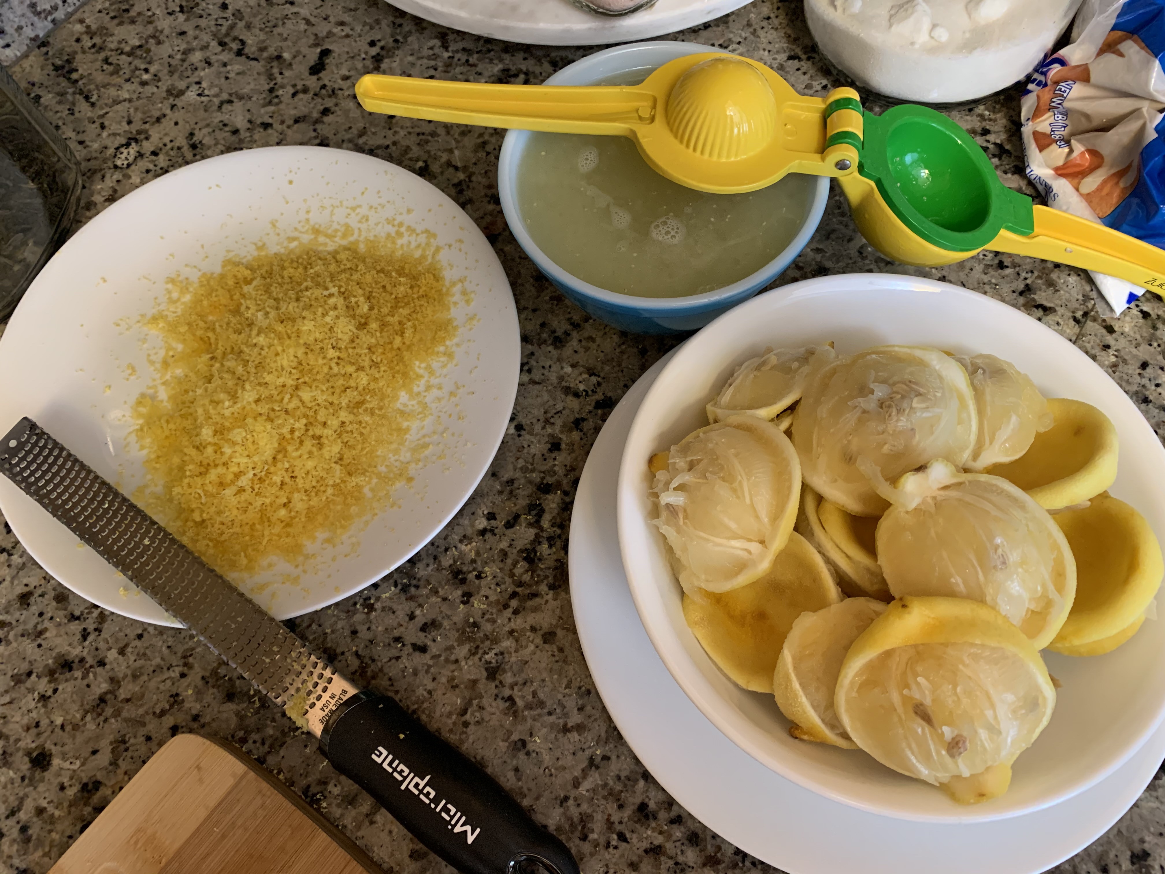 Aerial POV of lemon processing supplies: a dinner plate on the left with a layer of zest ready to flash freeze and a microplane rests across its bottom edge. A large bowl on the right containing zested and juiced husks, all pressed into similar dome shapes from the hand juicer. Centered at the top edge, a small bowl hold juice, and a yellow and green hand juicer lays across the top of the bowl.