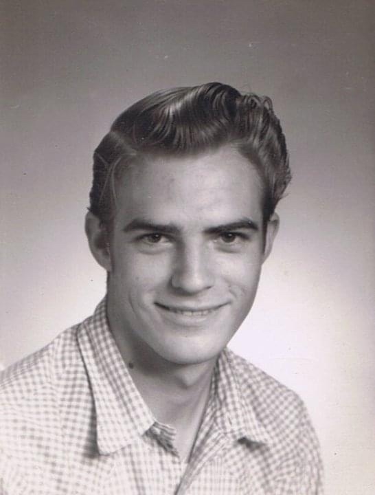 A black and white portrait of a fair skinned young man with a slender face and very short black hair, very slightly upstyled in a slight pompadour popular of the time. His black eyes twinkle and he has a slight smile, wearing a light colored, checkered, button down shirt.