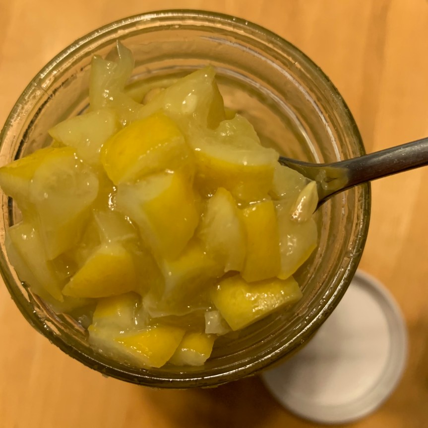Aerial POV of quart jar full of chopped, salt preserved lemons; a spoon, handle disappearing out of the top right corner of the frame, holds a scoopful of lemons above the surface of the jar’s contents.
