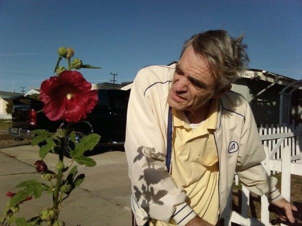 A large, red flower stands tall in the left half of this photo, Serena’s dad, much older and the deep lines characteristic of Parkinsons etched into his stubbly, salt and pepper face, stares at the flowerfrom the right side of the photo, stooped over slightly, His thin, salt and pepper hair flies a little loosely in the wind; he wears a white windbreaker and rests his hands on the handrests of a walker. The sky above is clear and blue.