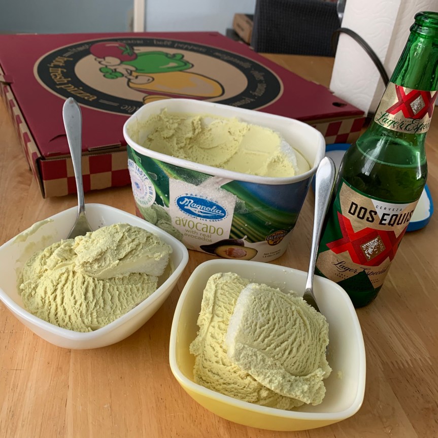 Two small, square bowls with avocado ice creamand spoons dominate the foreground of this picture, the open container of ice cream just behind them, and an open Dos Equis beer to the right of that. Behind the ice cream sits an unopen pizza box.