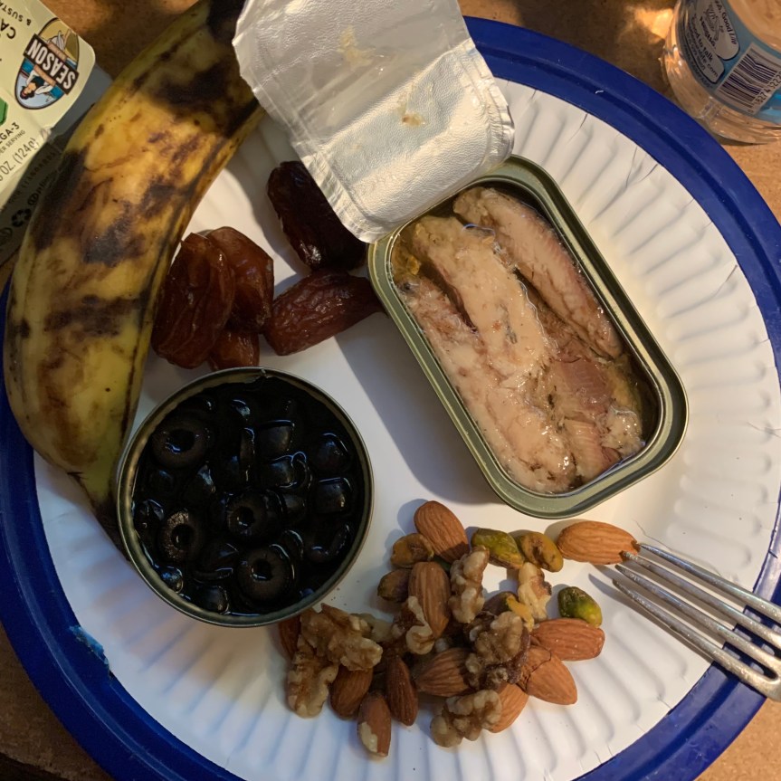 Tight aerial POV of a paper plate in a blue plastic plate holder. In the center, a small can of sliced black olives is open, and next to it, a rectangular tin of sardines is open with the top foil peeled back, but not fully removed. Below that, a small ppile of mixed nuts and above are a few dates, the top edge of the plate framed by a bruised and battered banana. The tines of a fork rest across the edge of the plate in the lower right corner.