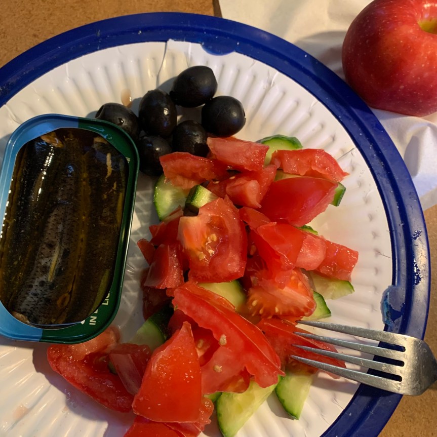 Aerial POV of a paper plate on a blue plastic plate tray, upon which sits an open tin of sardines and a few black olives on the left half, and a layered tomato cucumber salad that takes up most of the right side of the plate. An apple sits behind and to the right of the plate.