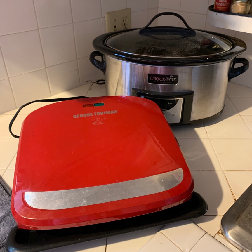 Close up of large slow cooker, back right, and red George Foreman grill, front left, sitting on a bare white tile countertop.