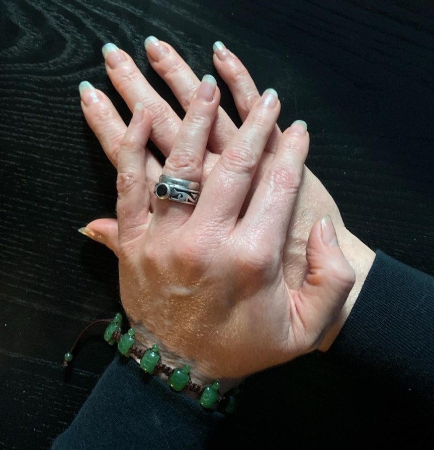 Pale feminine hands, left crossed at a slight angle on top of right, atop a black tabletop with a subtle wood grain and framed by black cuffs at the wrist. The left wrist holds a delicate bracelet of small carved jade turtles strung together with black nylon cord and the left ring finger is adorned with a heavy silver ring with ornate blackened carvings around the band and a large garnet and a simple thin silver band pushed on top of it. The nails are longer than sport length and not quite glamour length, uncolored and glossy with a simple clear coat of polish.
