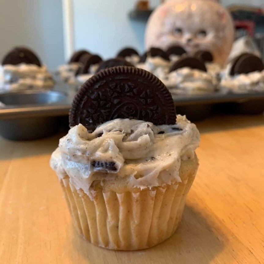 Extreme closeup side view of an Oreo cupcake with Oreo frosting and a half Oreo pressed decoratively into the frosting like a half cookie rising from the cupcake. The muffin tin containing the remainder of the batch is seen slightly out of focus in the background.