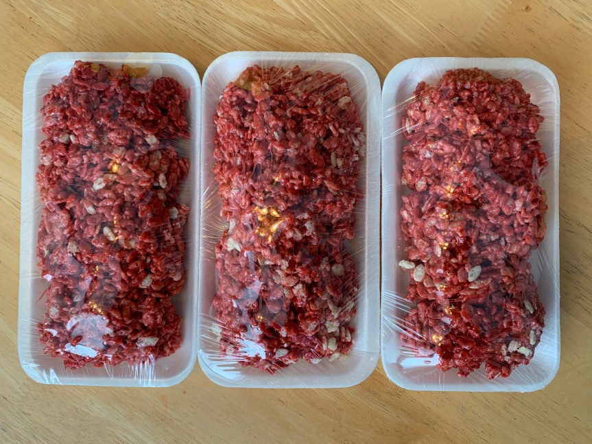 Landscape orientation aerial POV of three 1 lb. size styrofoam trays snuggled up side by side packed with realistic looking raw ground beef actually made from a rice krispy treat recipe, closed tight with cling wrap.
