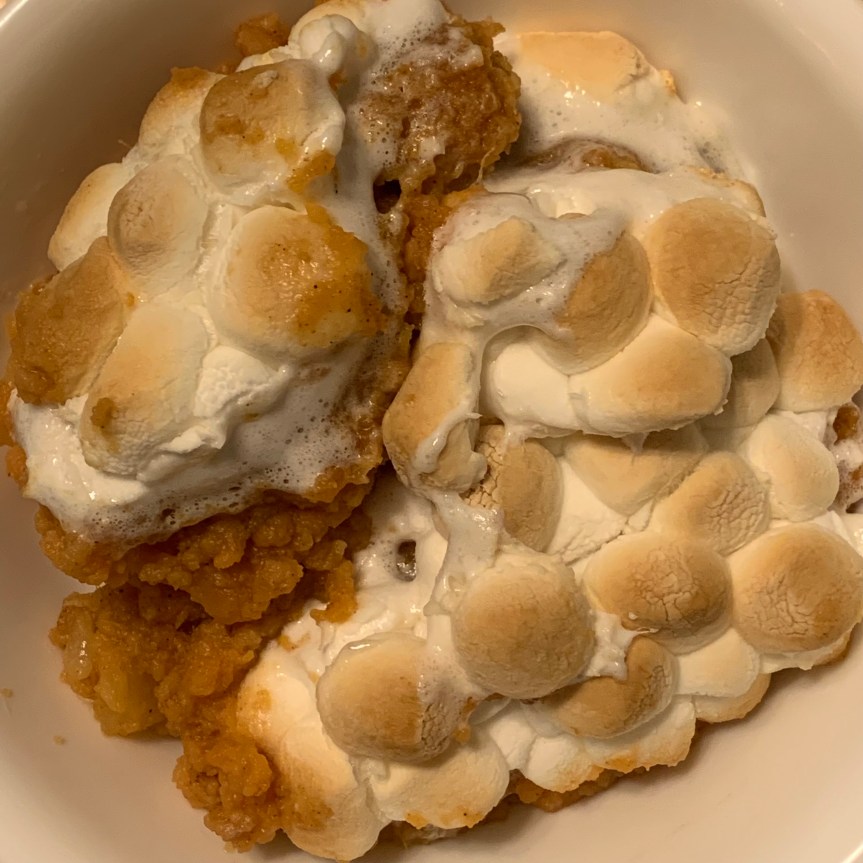 Close up aerial POV of a serving of yams and marshmallows in a white bowl. Rustic orange yams are seen peeking out from beneath pillowy clouds of golden toasted marshmallows.