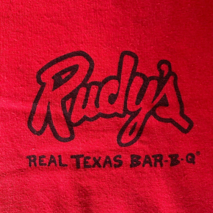 Close up of Rudy’s T-shirt pocket logo, the restaurant name in a stylized script with the words “real Texas barbecue“ underneath, black on red.