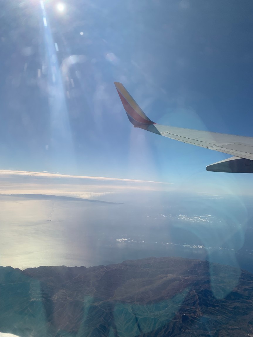 Portrait orientation view from an airplane window overlooking the California coast and Pacific Ocean. A bright ray of sunshine cuts down from the top of the picture and the tip of the airplane wing peeks into the top portion of the frame from the right.
