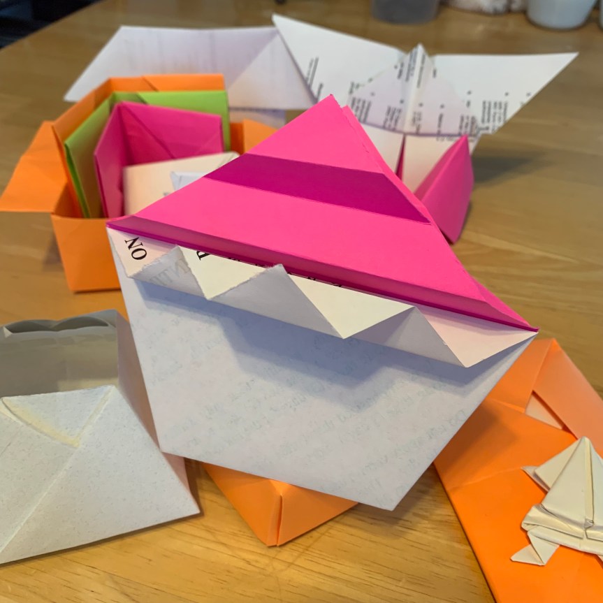 A large, whimsical origami cupcake sits atop an orange paper box. Various other origami models are seen around and behind, in white and bright, neon colors, a butterfly, envelopes and wallets, a little jumping frog, and others.
