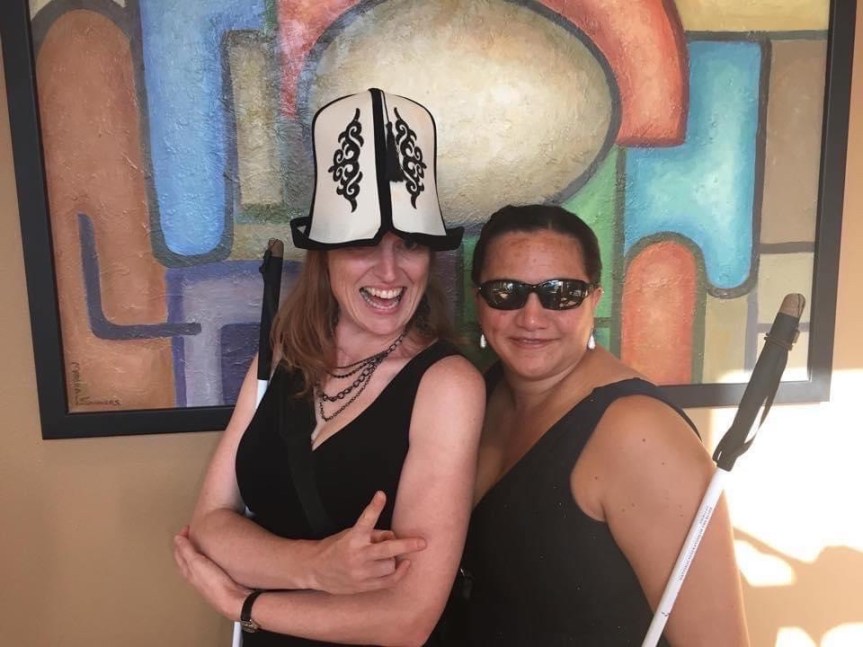 Serena, left, and LM, right, pause for a spontaneous photo in a hotel hallway in front of some colorful, abstract art; they are dressed a little fancy. Serena stands, arms folded and cane tucked into her right elbow, left shoulder out. She wears a traditional Kyrgyz kalpak, playfully lowered over her eyes and grins broadly. LM leans in, cane tucked into her left arm, wearing sunglasses and smiling lightly.