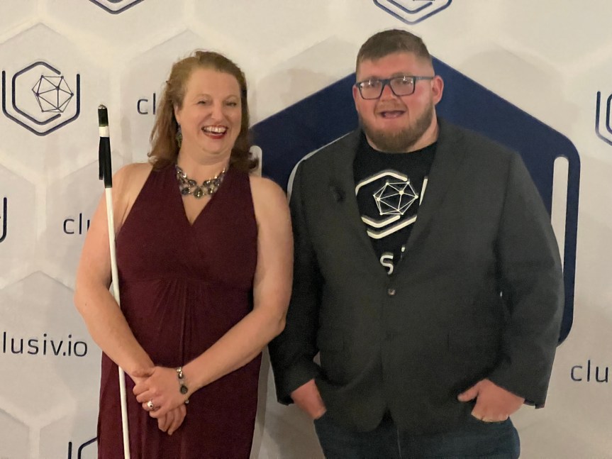 Blind Broad poses with Lukas Simianer in front of Clusiv‘s official media backdrop following the NFBC convention banquet. She is dressed in a long, burgundy evening dress with chunky, Gothic jewelry and light make up, smiling broadly. Lukas is dressed more casually, sporting a black Clusiv logo T-shirt and jacket.