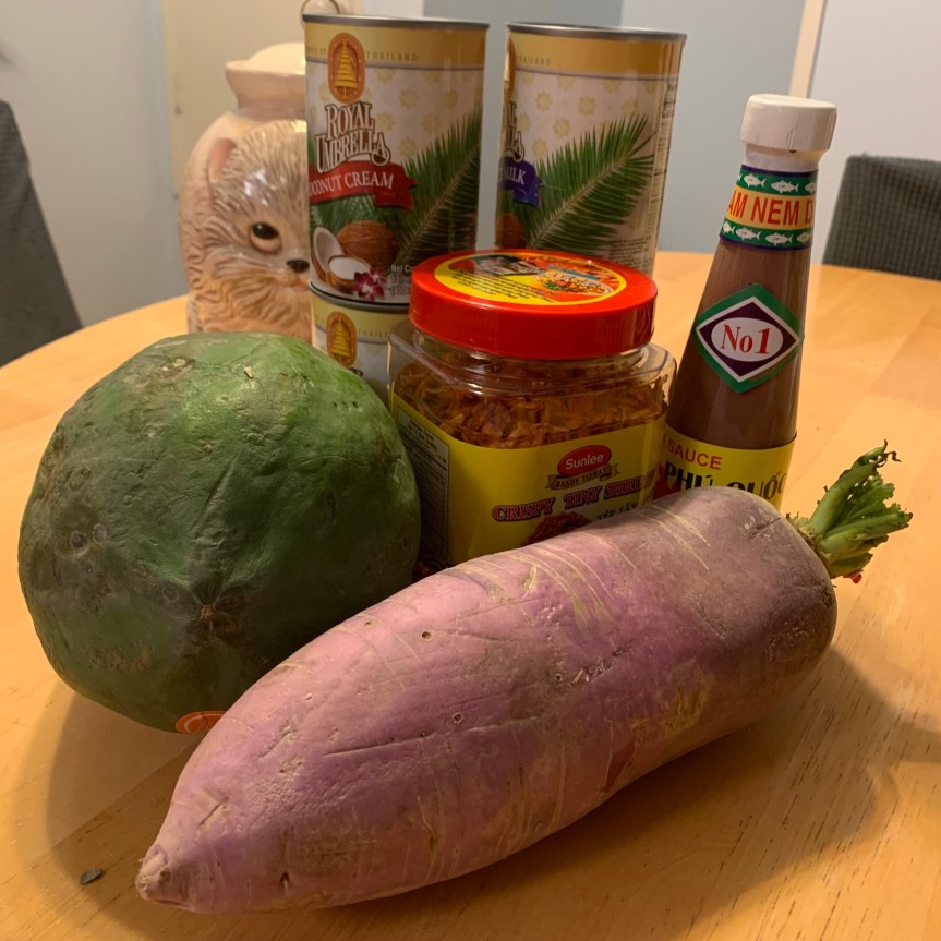 Two cans of coconut cream, two cans of coconut milk, a square plastic jar of tiny dried shrimp, and a small bottle of fish sauce stand on a table behind a green papaya and a purple daikon radish, resembling a large purple carrot.