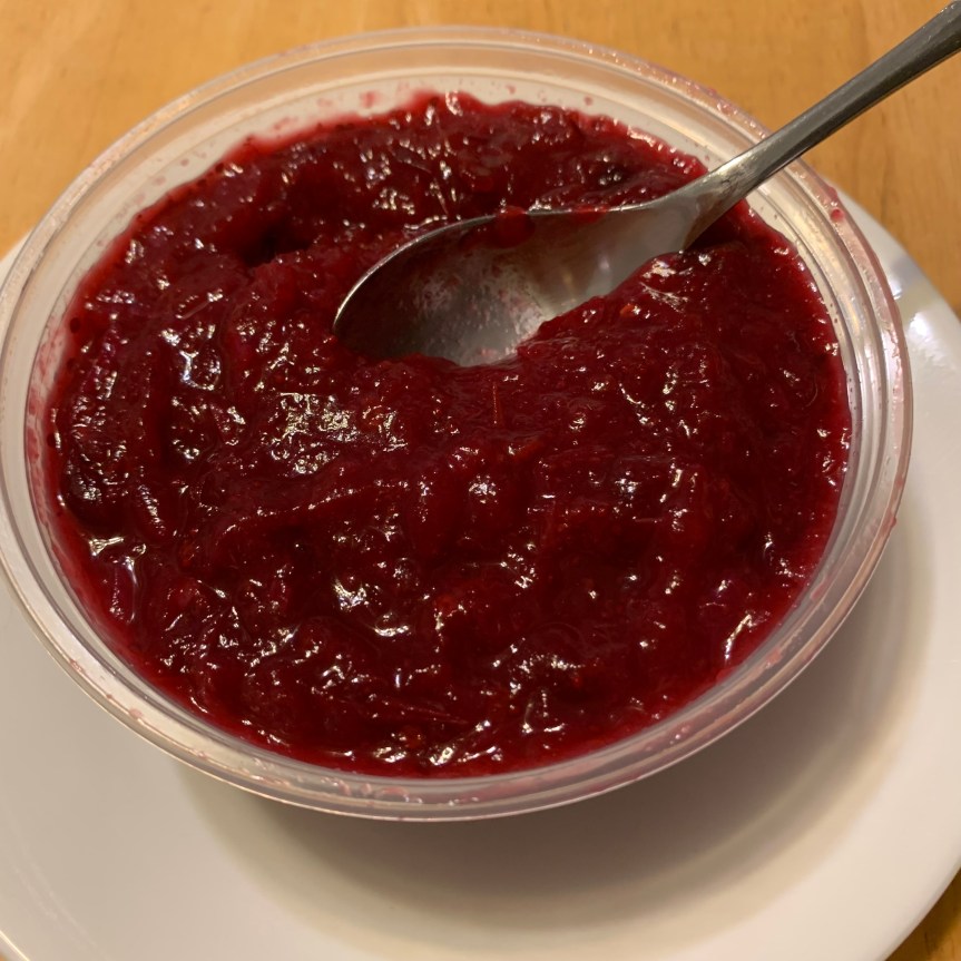 A plastic deli tub full of cranberry sauce with a spoon partially submerged into its surface sits on a white plate.
