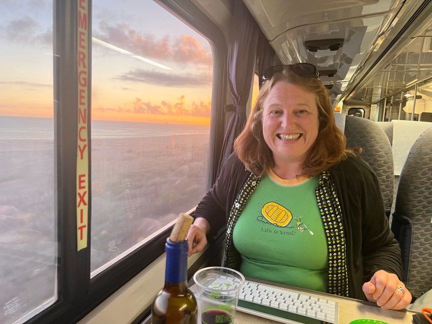 Blind Broad smiles broadly sitting at a table in a train car next to a wide window view of the sun setting over the Pacific Ocean. Her wireless keyboard is in front of her and a bottle of wine and other snacks line the table.