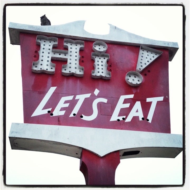 The sign portion of a large, kitchy street side, tall sign for Lompoc’s Hi! LET’S EAT restaurant. It is a faded dark red color with white letters and trim, Hi and its exclamation mark Large and prominent, let’s eat in all capital letters below. It has a 60s country aesthetic.