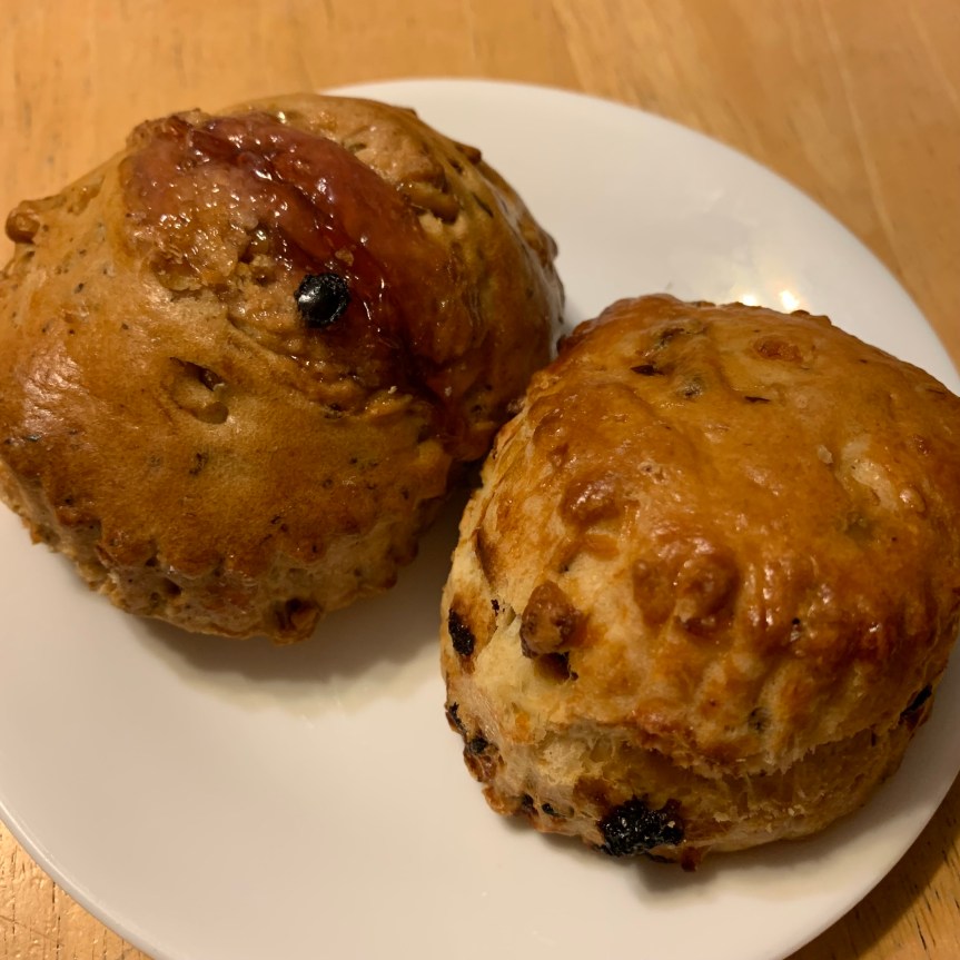 Two scones on a plate, round, not traditional wedge shaped, the left with a generous stripe of dark, gooey raspberry jam, the other flecked with small chunks of candied orange peel and dried currants.
