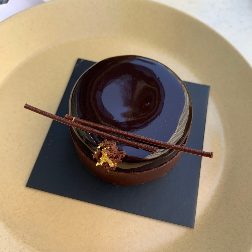 An individual sized Chocolate Imperial Cake, a tiny cake just a few bites big, with a chocolate mirror glaze and two wafer thin sticks of chocolate and a splash of edible gold garnish.
