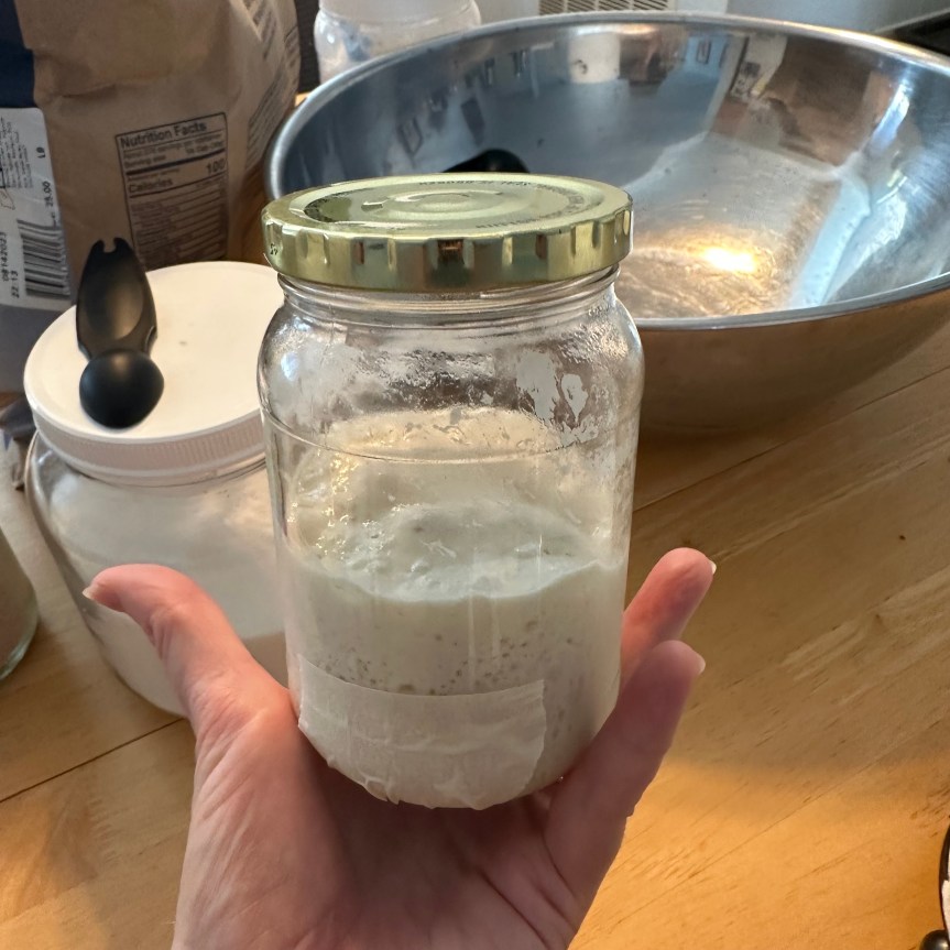 A hand holds up a small glass jar with a metal lid containing fluffy sourdough starter. A piece of masking tape on the outside of the jar marks the place where the starter began its rise, now doubled in size by the measure of the tape. A metal bowl, a jar of flour, and miscellaneous baking supplies are seen on the table in the background.