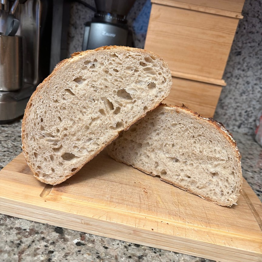 The picture shows two halves of a bread boule in cross-section placed on a wooden cutting board, one half resting partially atop the other. The bread has a golden-brown crust and a soft, airy interior with small holes. The cutting board is on a kitchen countertop, and in the background, there is a glimpse of a kitchen counter with a coffee grinder. The countertop has a speckled pattern. The bread looks freshly baked and appetizing. described with support from Be My AI.
