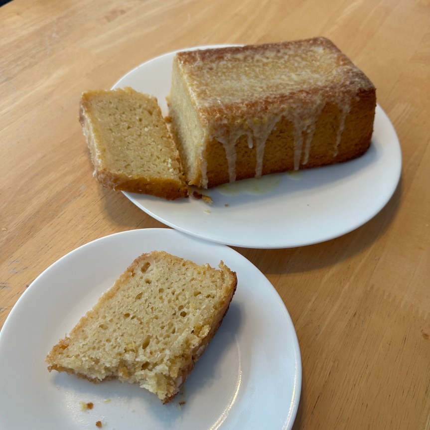 The picture shows a delicious looking loaf of cake on a wooden table. The cake is on a white plate and has a golden brown crust. It appears to be a lemon flavored cake with a drizzle of white icing on top. A slice has been cut from the loaf and is placed on a separate white plate in the foreground. The cake looks moist and has a soft, spongy texture. The background is simple, focusing on the cake, and the wooden table provides a warm, rustic feel. described with support from be my AI.