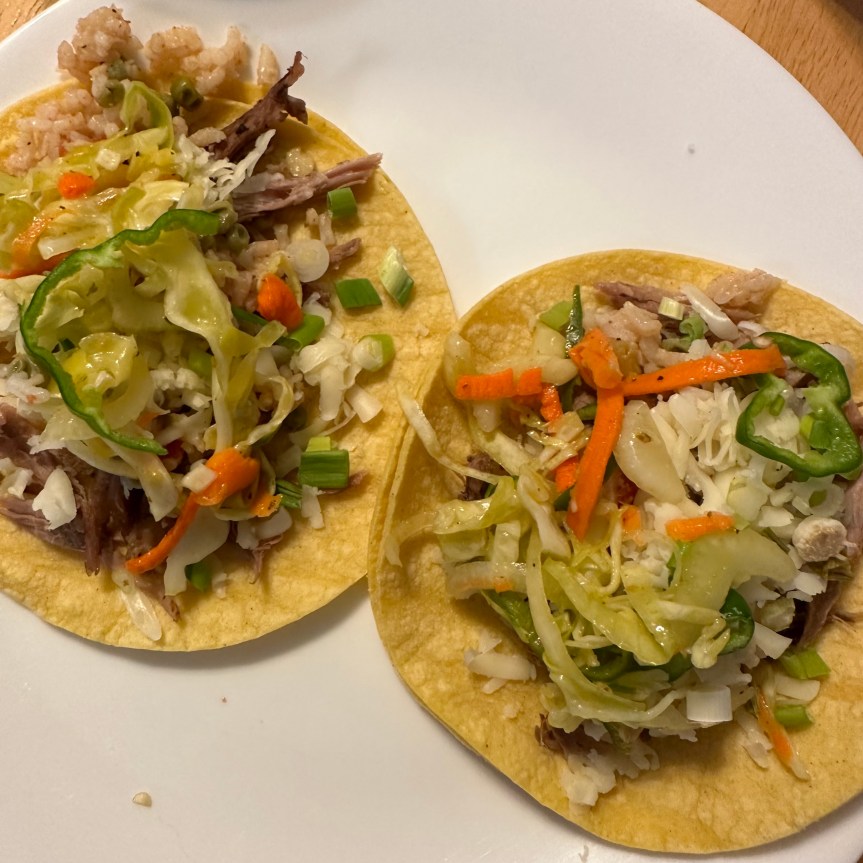 The picture shows two delicious tacos on a white plate. The tacos are on yellow corn tortillas and are generously filled with a mix of ingredients. There is shredded pork, and a colorful slaw made of shredded cabbage, carrots, and onion. There are also bits of chopped green onions and rice mixed in. The colors are vibrant, with the orange of the carrots, the green of the onions contrasting nicely with the yellow tortillas and the brownish meat. It looks like a fresh and appetizing meal. described with help from Be My AI.