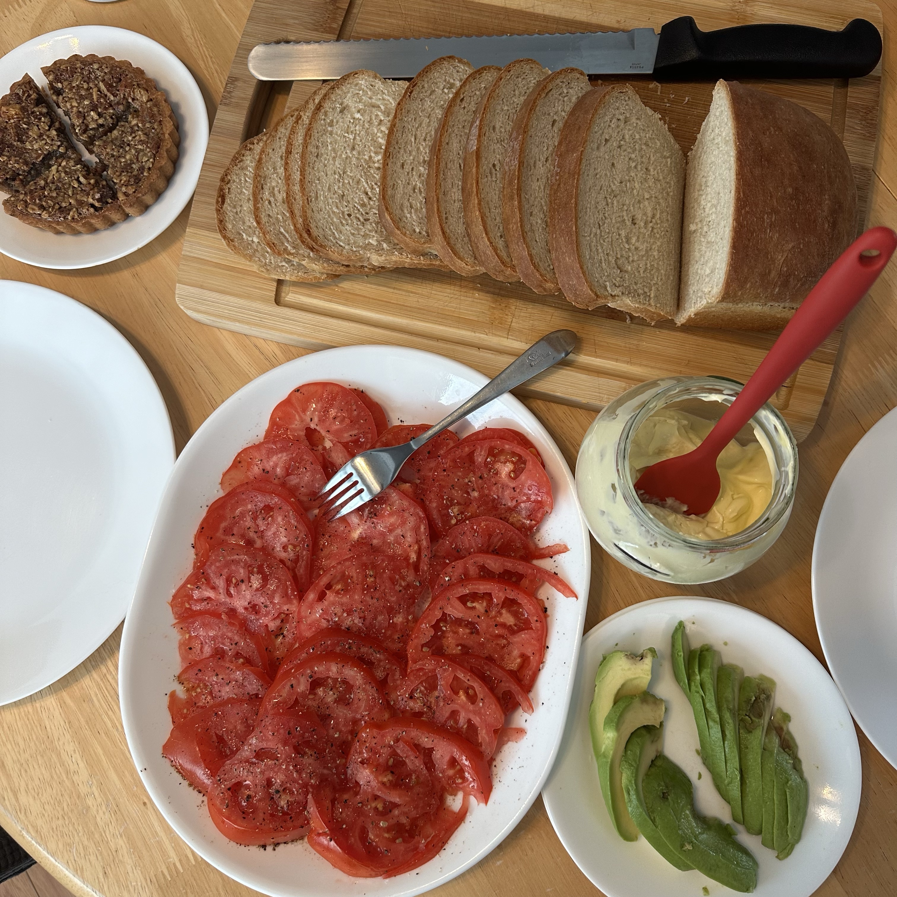 The image shows a spread of food items on a wooden table. There is a cutting board with a loaf of soft white bread that has been partially sliced, and a bread knife resting on the board. Below the cutting board, there is a white plate with neatly arranged slices of red tomatoes, seasoned with salt and pepper. To the right of the tomato plate, there is a jar of mayonnaise with a red spatula inside it. Next to the jar, there is a small white plate with slices of avocado. In the top left corner, there is a small round pecan tart that appears to be cut into four pieces. There are also a couple of empty white plates on the table. described with support from be my AI.