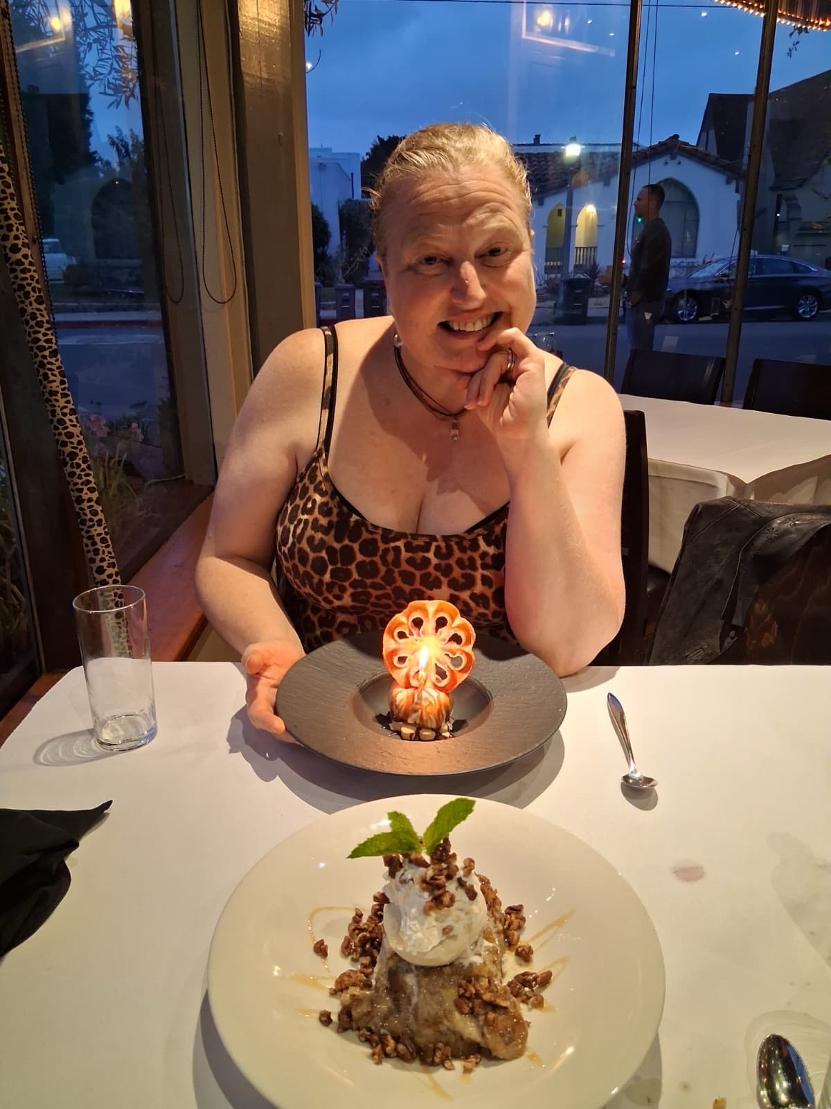 A person is seated at a dining table in a restaurant setting. The individual is wearing a leopard print top and has light-colored hair tied back. In front of them, on the table, is a dessert presented on a dark plate. The dessert features a decorative element resembling a flower with a lit candle in the center. To the right of the plate is a small spoon. In the foreground, closer to the camera, there is another dessert on a white plate. This dessert is topped with a scoop of ice cream, garnished with a sprig of mint, and sprinkled with what appears to be nuts or granola. The table is covered with a white tablecloth, and there is a clear glass on the left side of the table. The background shows large windows with a view of the street outside, where a car and some buildings are visible., July 20 Described using Be My AI.