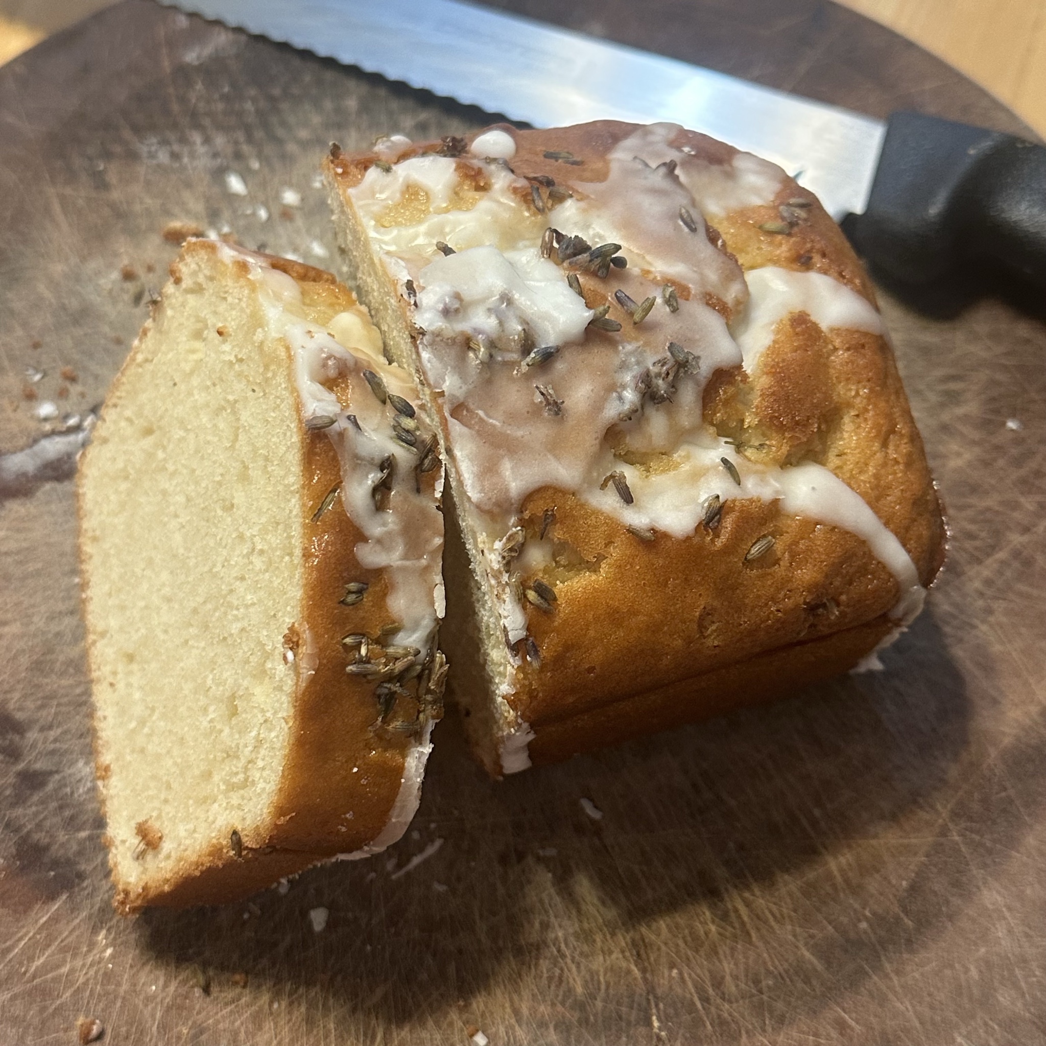 The image shows a loaf of bread on a wooden cutting board. The loaf is partially sliced, revealing a soft, light interior. The top of the bread is glazed with a white icing and sprinkled with small seeds or herbs. A serrated knife with a black handle is positioned nearby on the board. The bread appears to be freshly baked, with a golden-brown crust., Halcyon-Foothill, San Leandro, CA, 8:10 PM described using B my AI.